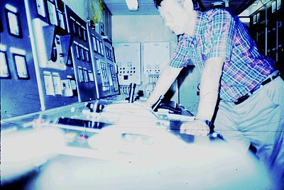 Vintage industrial control room with operator monitoring analog gauges and switches, likely from the 1960s-1970s. Blue-tinted...