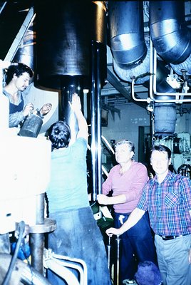 Industrial facility interior featuring four workers in casual 1970s attire. Two men in foreground—one in plaid shirt, another...