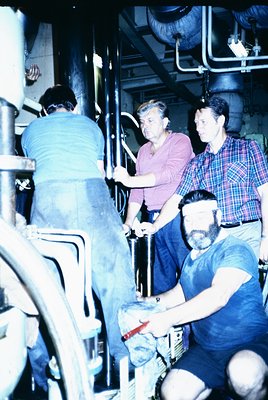 Industrial setting featuring four workers in a dimly lit, utilitarian space with exposed pipes and machinery. Three men in ca...