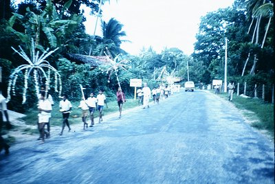 Vintage street scene with a group of uniformed individuals marching in formation along a rural road lined with tropical folia...
