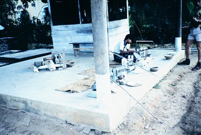 Vintage outdoor workspace with concrete slab and wooden supports, likely a 1970s–1980s workshop or artisan studio. Two indivi...