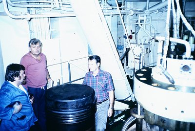 Industrial facility interior featuring three men in mid-20th-century workwear. One wears a blue jumpsuit, another a plaid shi...