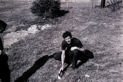 Black-and-white candid of a man kneeling on grassy terrain, holding a small object in hand. His posture suggests focus on the...