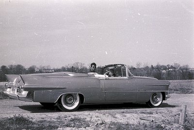 Classic 1950s convertible with tailfins parked in rural setting. Two figures seated inside, one driver, one passenger. Distin...