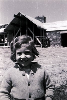 Vintage black-and-white photo of a smiling child in a buttoned cardigan, standing outdoors near a rustic stone building with ...