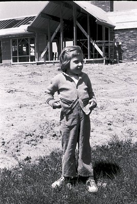 Young girl in early 1960s-style overalls poses outdoors near a brick building with a sloped roof. She holds a small object in...