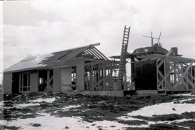 Mid-20th century construction site featuring skeletal wooden frame of a single-story building with gabled roof. Adjacent is a...