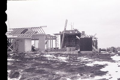 Mid-20th century industrial construction site by waterfront, featuring skeletal concrete structures and scaffolding. Snow cov...