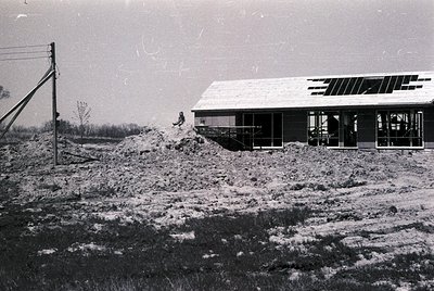 Mid-century modern building under construction, surrounded by excavated earth. Single-story structure with flat roof and larg...