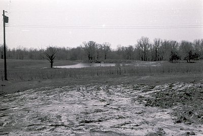 Rural landscape with plowed field and sparse winter trees, likely late 1940s–1960s. Snow-covered ground and barren branches s...