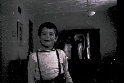 Vintage black-and-white portrait of a young boy in suspenders, smiling directly at the camera. Indoor setting with blurred re...