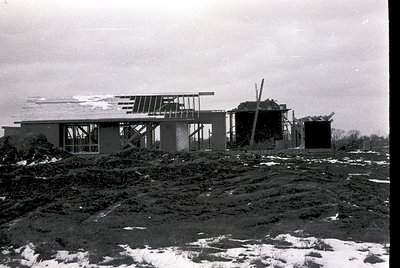 Mid-century modern construction site featuring a partially built concrete home with large glass windows and exposed structura...