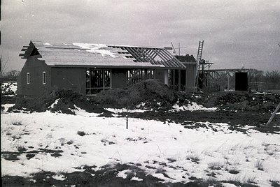 Single-story brick house under construction, winter 1950s–1960s. Exposed framing, partial roofing, and scaffolding indicate m...