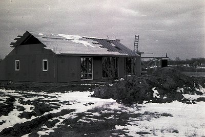 Single-story prefab building with gabled roof, partially snow-covered ground, and industrial scaffolding in background. Likel...