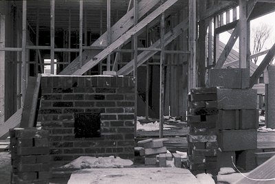 Mid-construction brick fireplace framed by wooden scaffolding; stacked bricks and lumber indicate early-stage masonry. Likely...