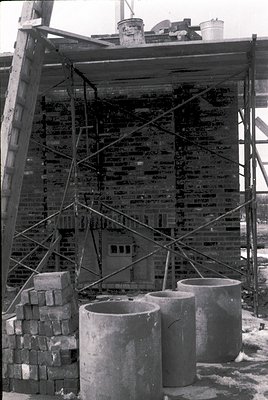 Mid-20th century brick construction site with scaffolding and concrete mixing barrels. Stacked bricks and paint cans atop sca...
