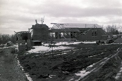 Mid-century modern building under construction, skeletal frame with exposed beams and scaffolding. Snow patches indicate wint...