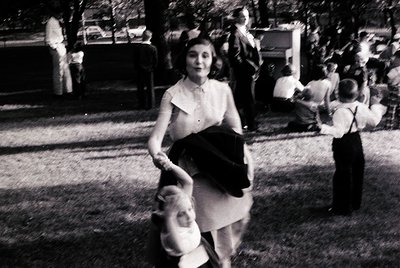 Vintage black-and-white photo of a woman mid-dance with a child in mid-20th century outdoor setting. Woman wears a knee-lengt...