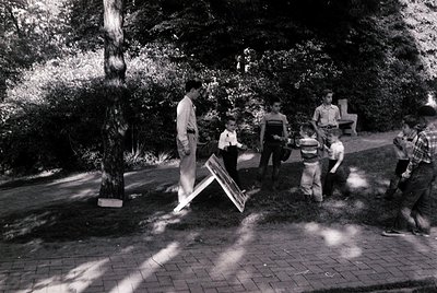 Group of children in 1960s-70s park setting, carrying a large wooden structure. Lush greenery and paved paths visible. Casual...