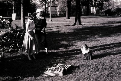 Mid-20th century park scene: woman in structured skirt and hat supervises toddler playing with a checkered board on grass. Su...