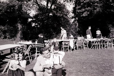 Mid-20th century outdoor gathering under dense trees, featuring a long picnic table setup. Foreground shows a man in a light ...