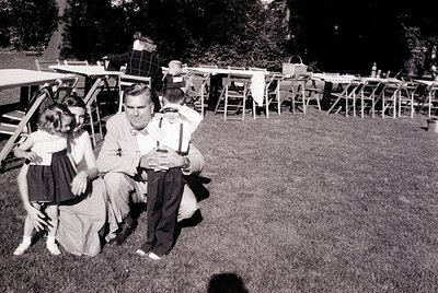 Mid-century outdoor gathering in a lush garden setting, featuring a man in a suit and tie seated with a young girl on his lap...