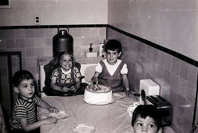 Mid-century kitchen scene with three children (two boys, one girl) gathered around a round birthday cake on a tiled table. Wh...