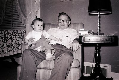 Mid-20th century indoor portrait: man in glasses and striped shirt cradles a toddler in a patterned armchair beside a vintage...