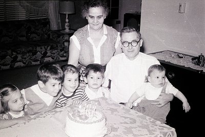 Family portrait from the 1960s–70s, likely in a mid-century American home. Five children and two adults pose around a birthda...