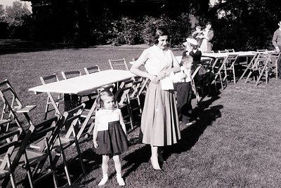 Mid-20th century outdoor gathering in a lush garden setting. A woman in a knee-length dress and a young girl in a matching dr...