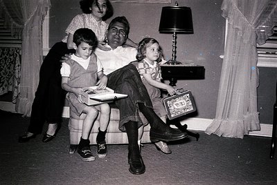 Vintage black-and-white family portrait in a mid-century living room. Three children and two adults pose on a sofa, surrounde...