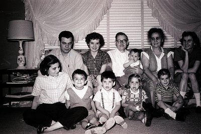 Family portrait from the 1960s, likely in a mid-century American home. Eight adults and five children pose indoors with vinta...