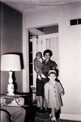A 1950s-1960s indoor family portrait: woman in a plaid blouse and skirt holds a baby, while a young boy in a coat and cap sta...