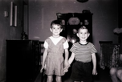 Two young boys pose indoors in mid-20th century attire—striped shirt and shorts, V-neck shirt with shorts. Wooden cabinet wit...