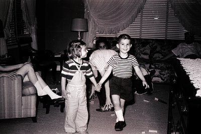 Mid-20th century indoor family moment: two children in striped shirts and overalls playing in a dimly lit living room with a ...
