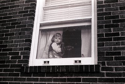 Black-and-white photo of a child peering through a window framed by brickwork. The child wears a patterned dress and holds a ...