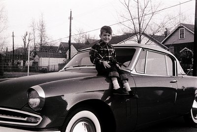 Vintage black-and-white photo of a boy perched atop a classic sedan, likely a 1950s–1960s American model. Suburban residentia...