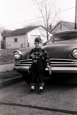 Mid-20th-century boy poses on vintage car hood, holding a small object. Classic suburban neighborhood with mid-century reside...