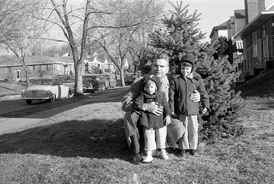 Three people—an adult man and two children—pose outdoors on grass beside a residential street with parked vintage cars (1950s...