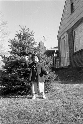 A black and white photograph of a small child standing in a grassy yard. The child wears a dark coat, a hat, and light-colore...