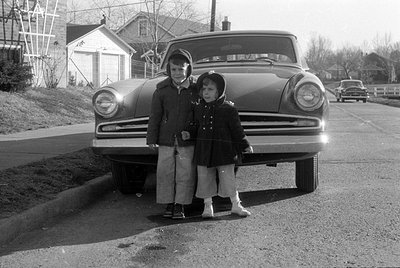 Two children in winter coats and hoods stand in front of a vintage car parked on a residential street. A house and a second c...