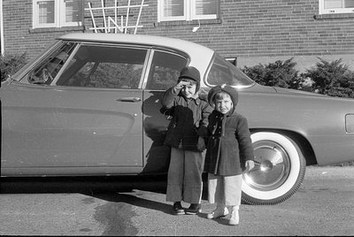 Two children pose beside a vintage car in a residential driveway, waving at the camera. The car has whitewall tires and a cla...