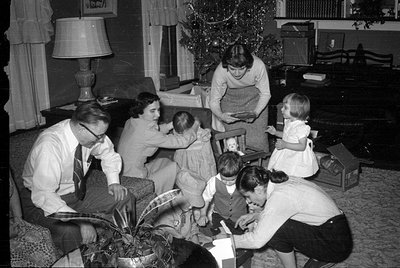 A black and white indoor photograph of six people gathered around a decorated Christmas tree. A man, three women, and two chi...