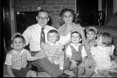 A black and white photograph of a man, a woman, and four young children posed in a living room. The man wears glasses and a t...