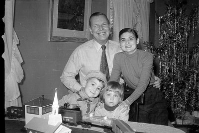 A black and white photograph of a man, a woman, and two children posing behind a model train set and a small white church mod...