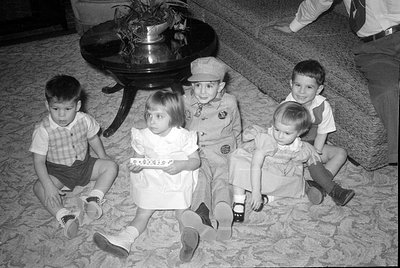 Four young children sit on a patterned carpet next to a dark wooden table. One child wears a striped uniform with a matching ...