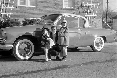 A black and white photograph of a man and two children posing beside a coupe with whitewall tires. They are positioned in fro...