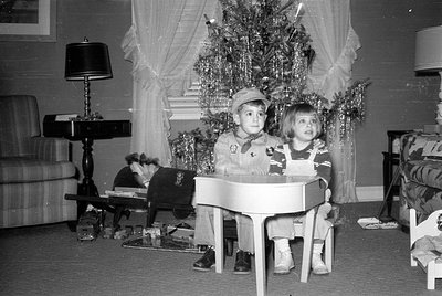 Two young children sit behind a small white toy piano in a living room. A decorated Christmas tree with tinsel stands behind ...