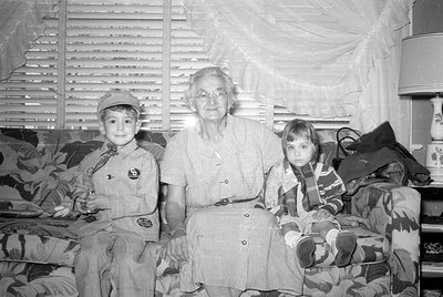 A black and white photograph of an older woman and two young children sitting on a patterned sofa. The child on the left wear...