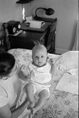 A black and white photograph of an infant sitting on a patterned bedspread. A woman is partially visible on the left, holding...
